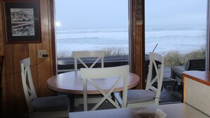 Un interior de madera con una mesa redonda y sillas blancas, con vistas a una playa y olas.