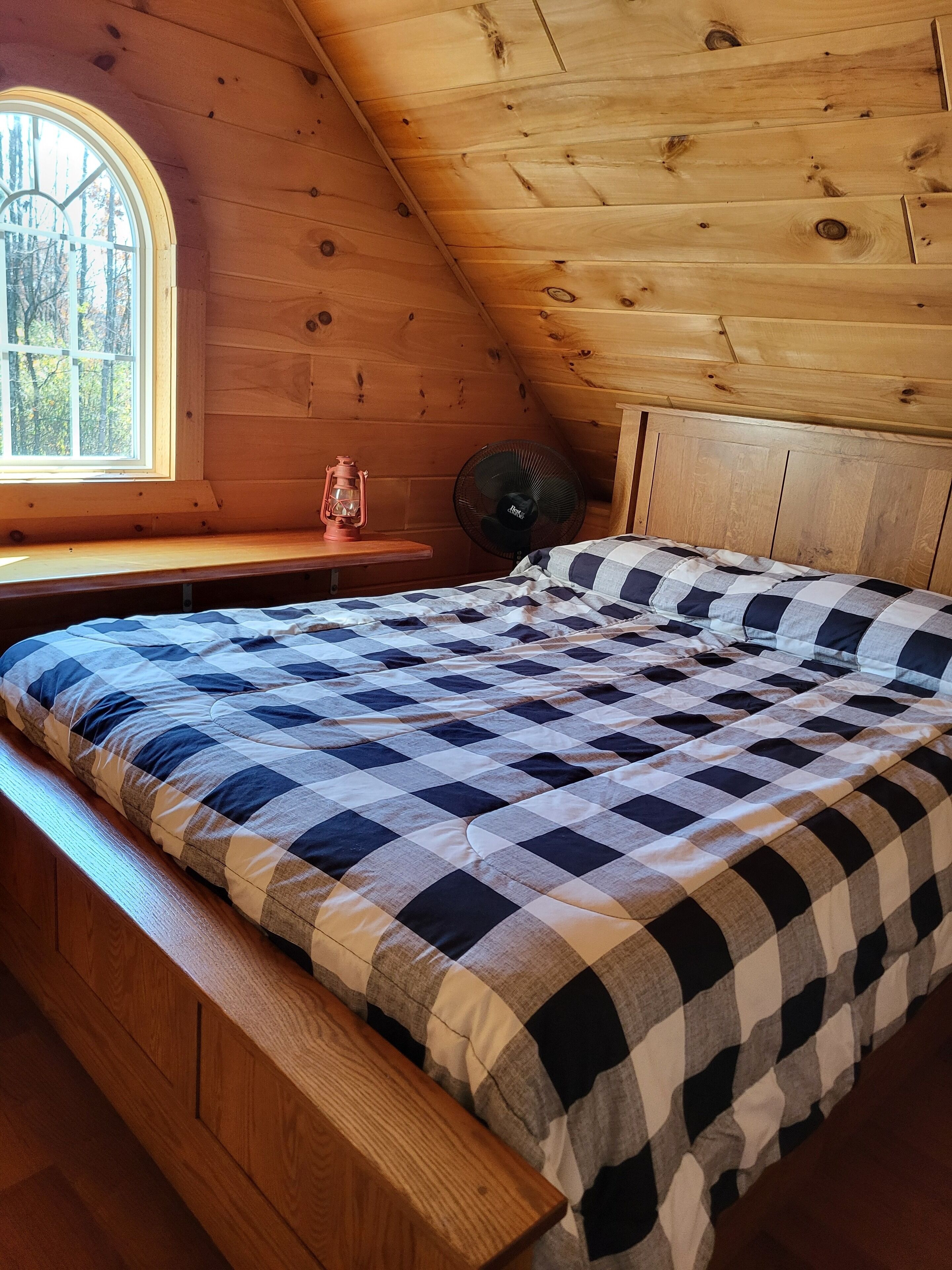 A cozy attic bedroom with a checkered bedspread, a wooden bench, a small table with a lantern, and a fan.
