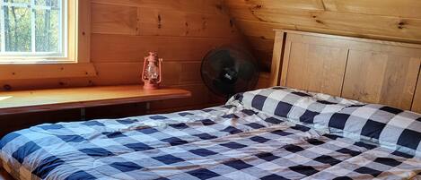 A cozy attic bedroom with a checkered bedspread, a wooden bench, a small table with a lantern, and a fan.