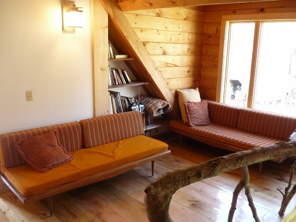 A cozy cabin interior with two striped sofas, a wooden bookshelf, and a large window.