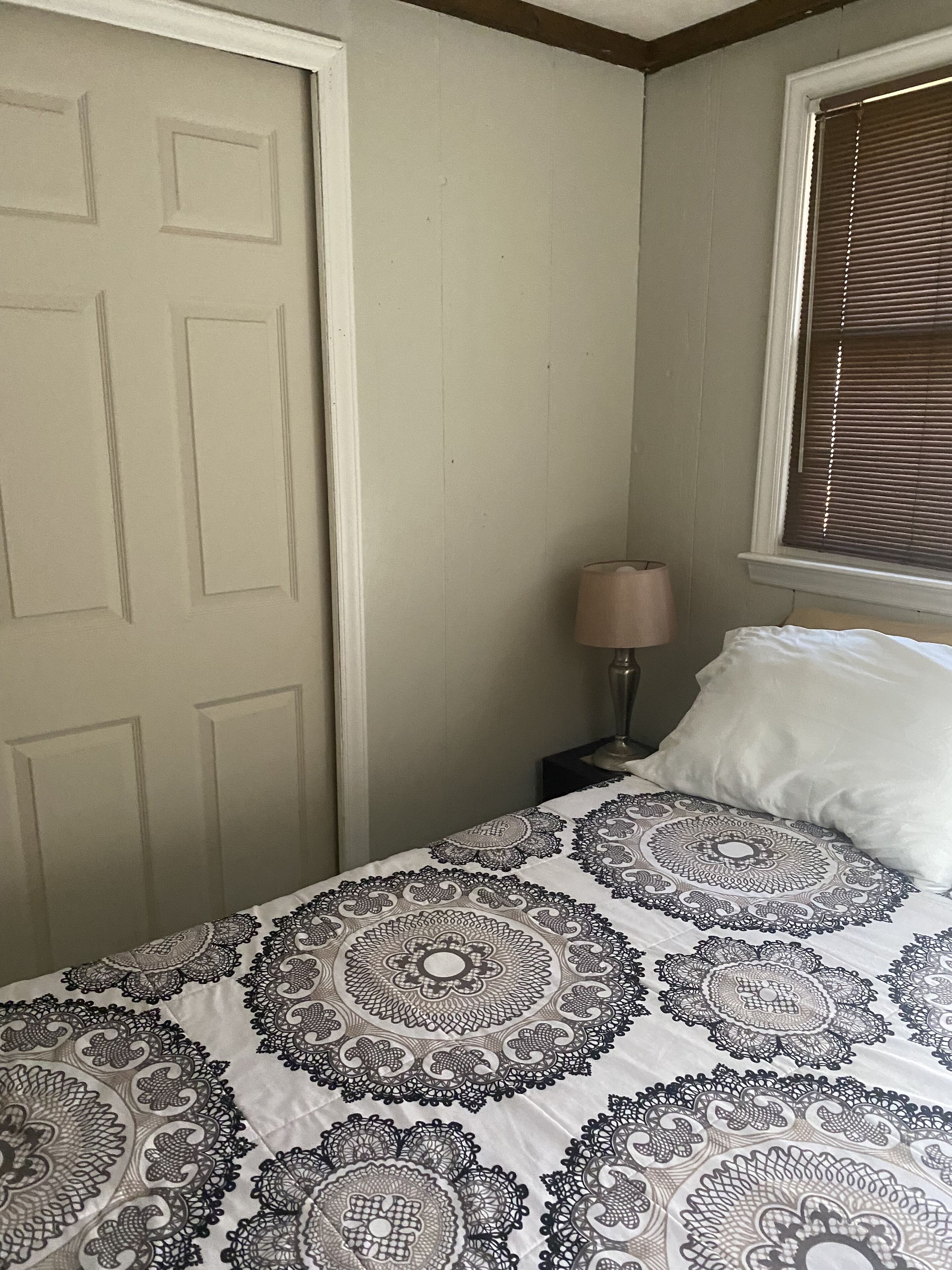 A bedroom with a bed, a nightstand, a lamp, a window with blinds, and a door.