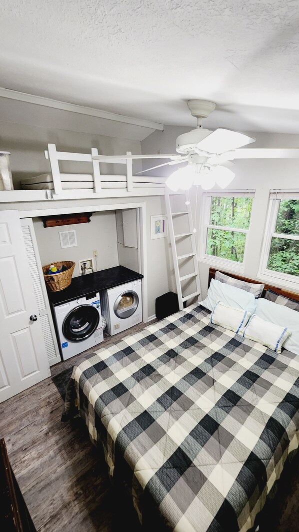 A compact room with a bunk bed, a washing machine, a ceiling fan, and a window with a view of greenery.