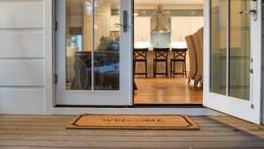 A wooden deck with a welcome mat, a glass door leading to a kitchen with a dining area.