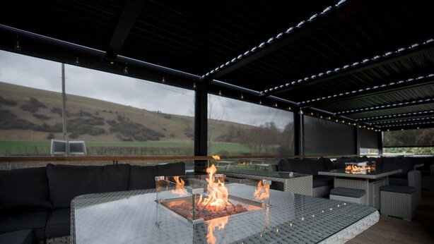 An outdoor seating area with a glass-top table and a central fire feature. - Tottington Manor (Henfield)