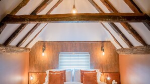 A bedroom with a sloped ceiling, wooden beams, a large bed with red bedding, two bedside lamps, and a window with blinds. - Tottington Manor (Henfield)