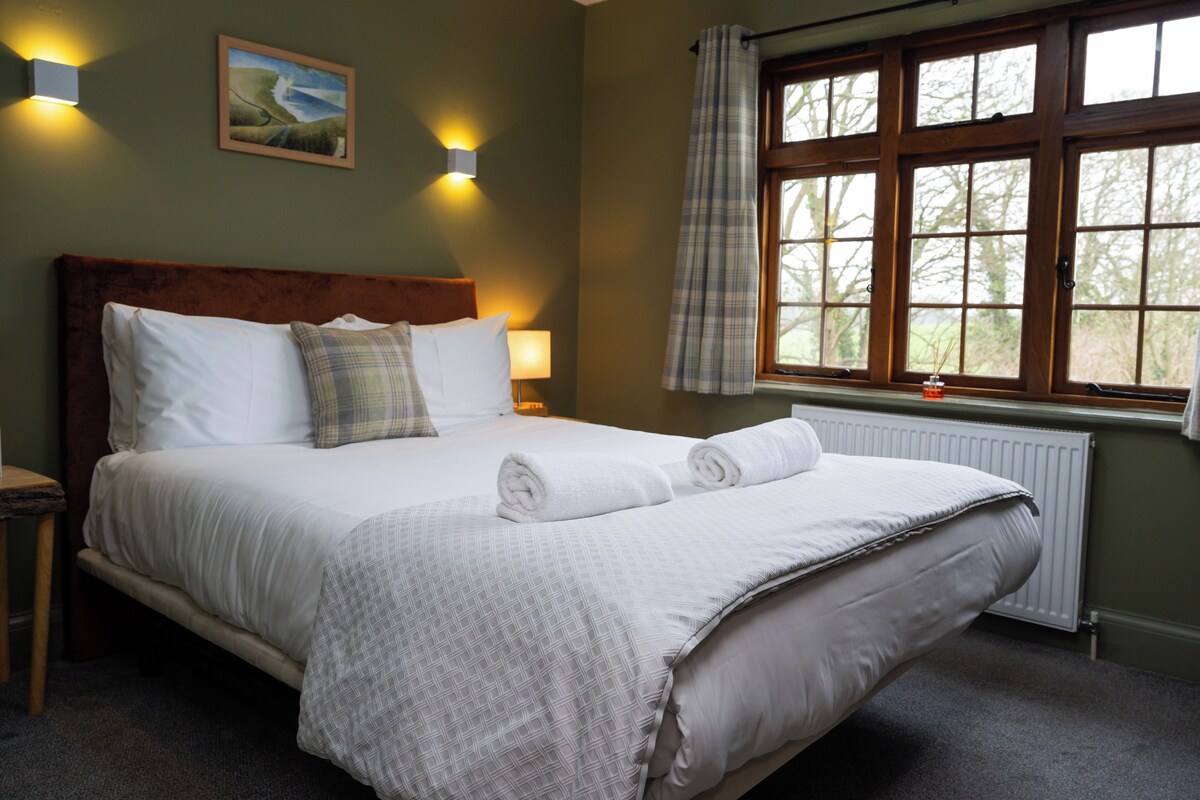 A neatly made bed with white linens and a plaid blanket, a wooden headboard, and a window with a view of trees.