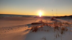 Een strand bij zonsondergang met zand, schaarse begroeiing en een hek in de verte.