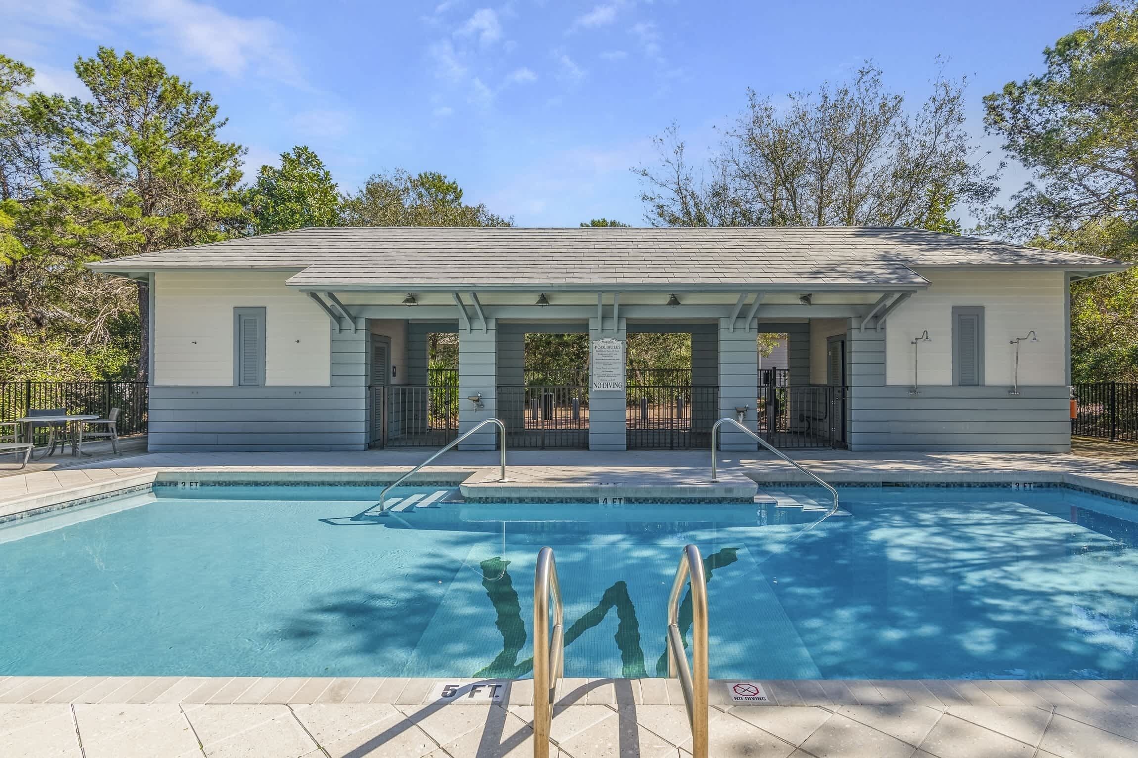 A rectangular swimming pool with a covered deck area, surrounded by trees and a clear sky.