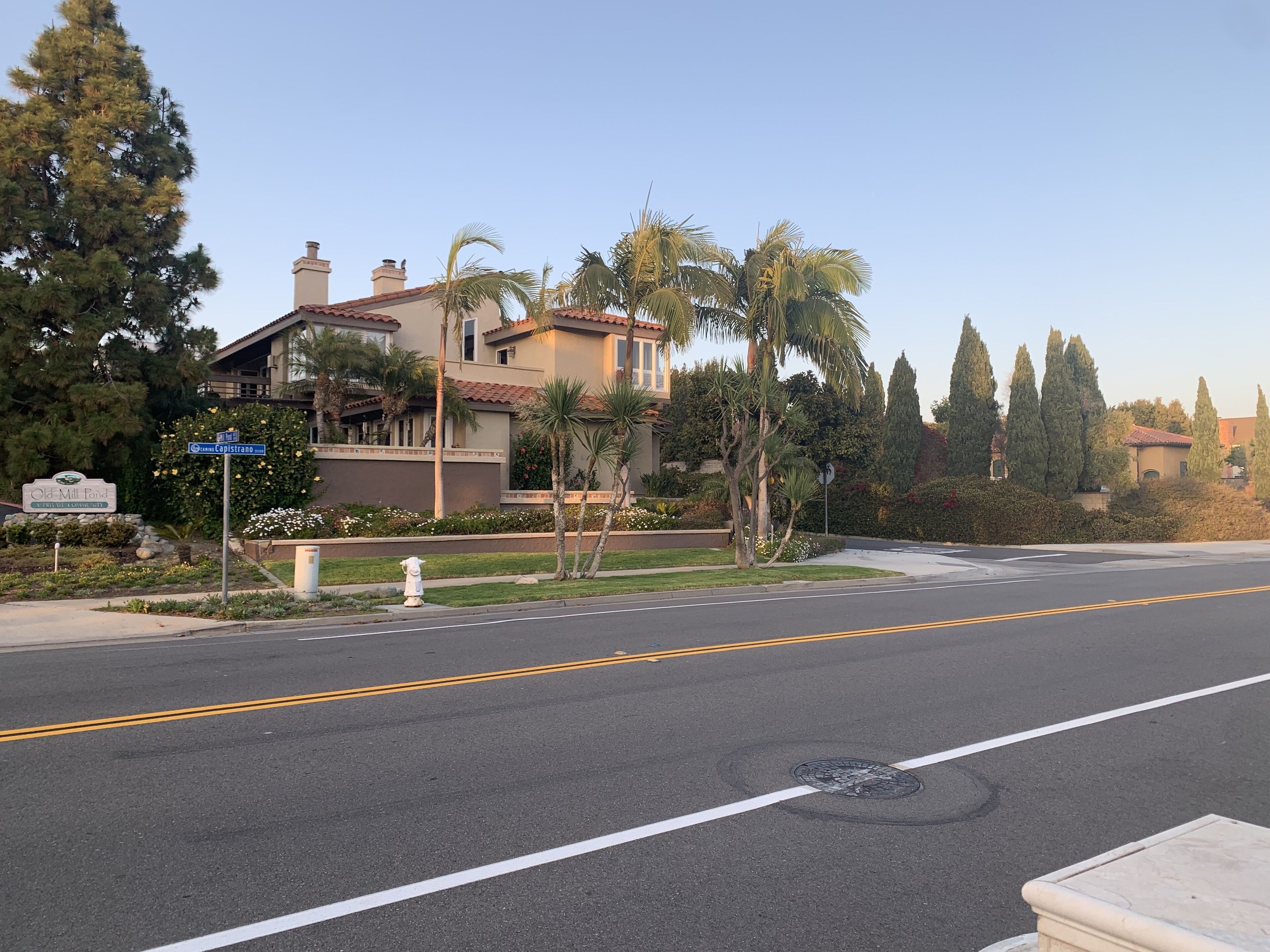 Una calle residencial con una casa de dos pisos, palmeras y cielo despejado.