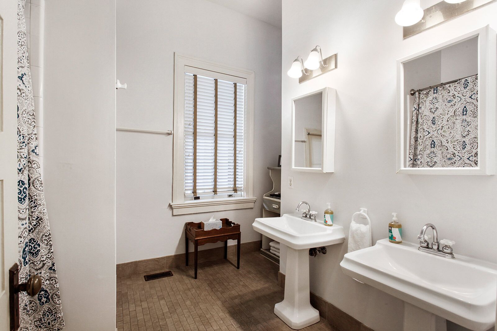 Un baño con dos lavamanos, una ventana con persianas, un espejo y un perchero para toallas.