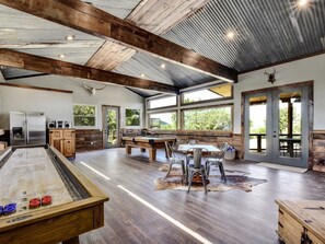 Un grand espace intérieur comprenant une table de billard, une table à manger et des chaises. De grandes fenêtres laissent entrer la lumière naturelle et offrent une vue sur la verdure environnante.