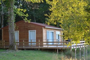 A wooden cabin with a deck, surrounded by trees and grass. - Les chalets de la héronnière (Chef-Boutonne)