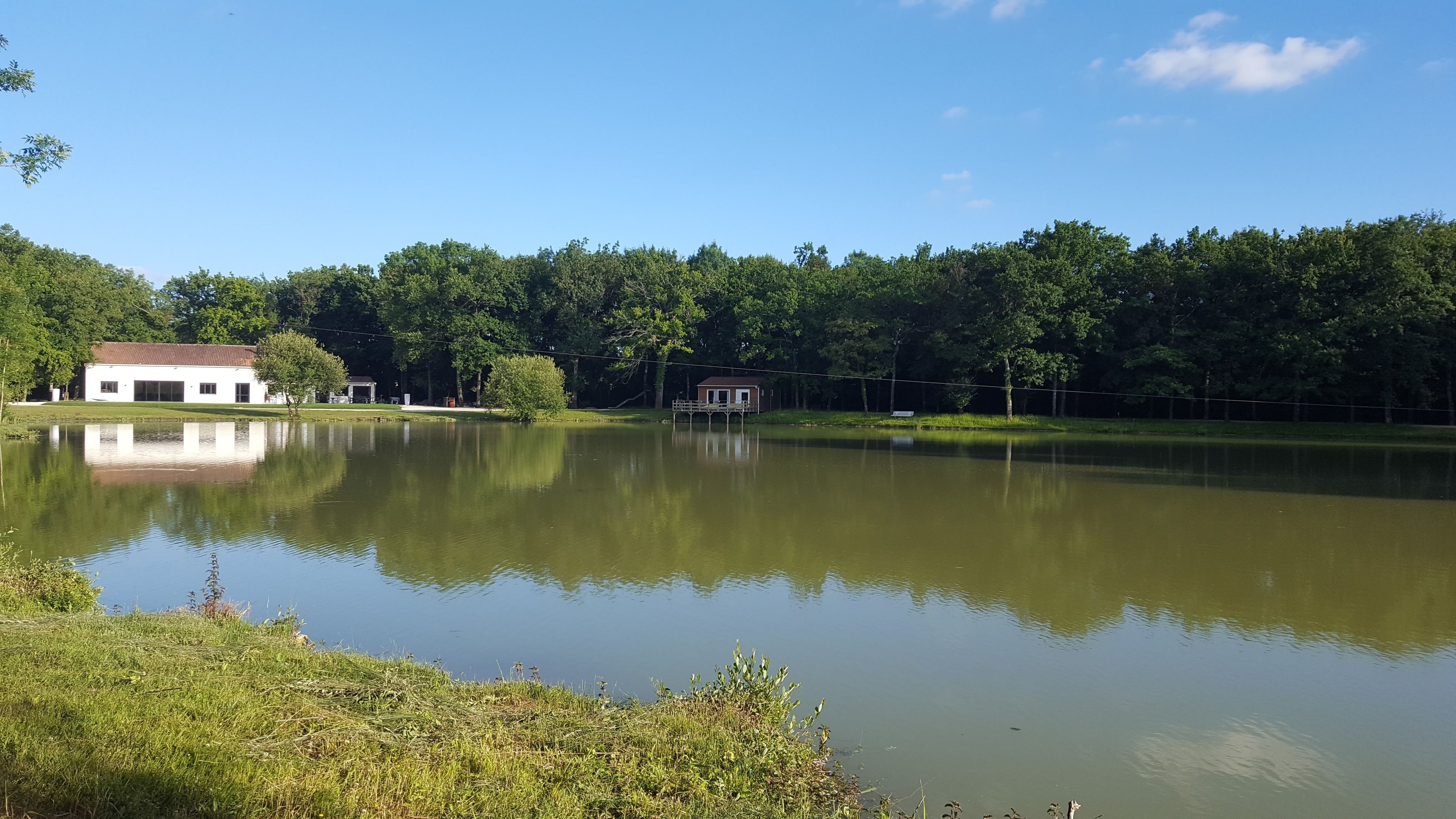 Un lac paisible, avec un petit quai et, au loin, un bâtiment blanc surmonté d’un toit rouge.