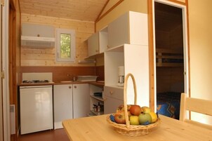 Une petite cuisine équipée d’un réfrigérateur, d’un évier et d’une fenêtre. Un panier de fruits est posé sur la table.