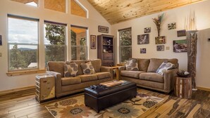 A spacious living room with two sofas, a coffee table, and a wooden cabinet. Large windows provide natural light and views of the outdoors. - Custom Home with Two Private Master Suites and Heated Garage (Angel Fire)