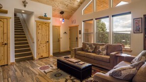 A spacious living room with a high ceiling, wooden floors, a staircase, and large windows offering a view of the outdoors. - Custom Home with Two Private Master Suites and Heated Garage (Angel Fire)