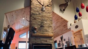 A cozy living room with a stone fireplace, wooden beams, and a deer head decoration. - Custom Home with Two Private Master Suites and Heated Garage (Angel Fire)