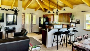 A spacious living area with a high ceiling, wooden beams, a kitchen with a central island, and a dining area with bar stools. - Nice, Big House near Kehena/ Pohoiki Black Sand Beaches and Lava Fields. (Pahoa/Kalapana-Seaview)