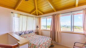 A bedroom with a bed, a desk, and a chair. There are windows with curtains, a ceiling fan, and a view of the outdoors. - Nice, Big House near Kehena/ Pohoiki Black Sand Beaches and Lava Fields. (Pahoa/Kalapana-Seaview)