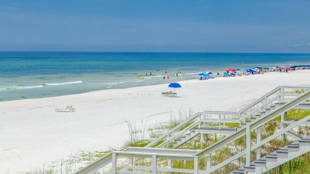 Ein Strand mit weißem Sand, eine Treppe, die zum Ufer hinabführt, und ein klarer, blauer Himmel.
