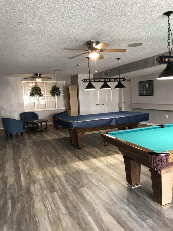 A pool table with a blue cloth, a ceiling fan, and pendant lights in a room with wooden flooring and a white ceiling. - Cozy & Mod condo, Walk to Beach, Self check In-Out, WiFi, 2 Heated Pools, Piers+ (Madeira Beach)