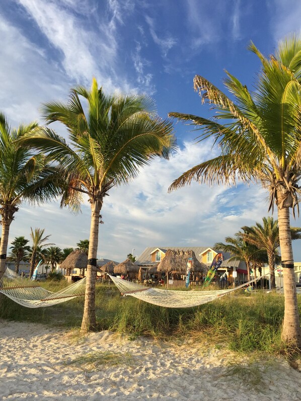 A beach scene with palm trees, a hammock, and a building with thatched roofs. - Cozy & Mod condo, Walk to Beach, Self check In-Out, WiFi, 2 Heated Pools, Piers+ (Madeira Beach)