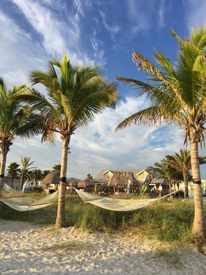 A beach scene with palm trees, a hammock, and a building with thatched roofs. - Cozy & Mod condo, Walk to Beach, Self check In-Out, WiFi, 2 Heated Pools, Piers+ (Madeira Beach)