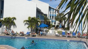 Une piscine entourée de palmiers et bordée d’un bâtiment moderne, avec des chaises longues et des parasols à proximité.