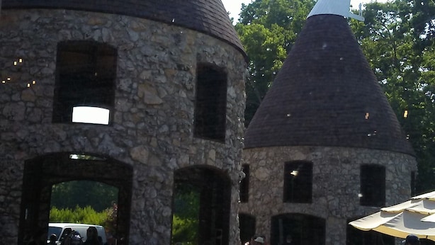 Un edificio de piedra con tejados cónicos, terraza al aire libre con mesas y sillas, y personas cenando.