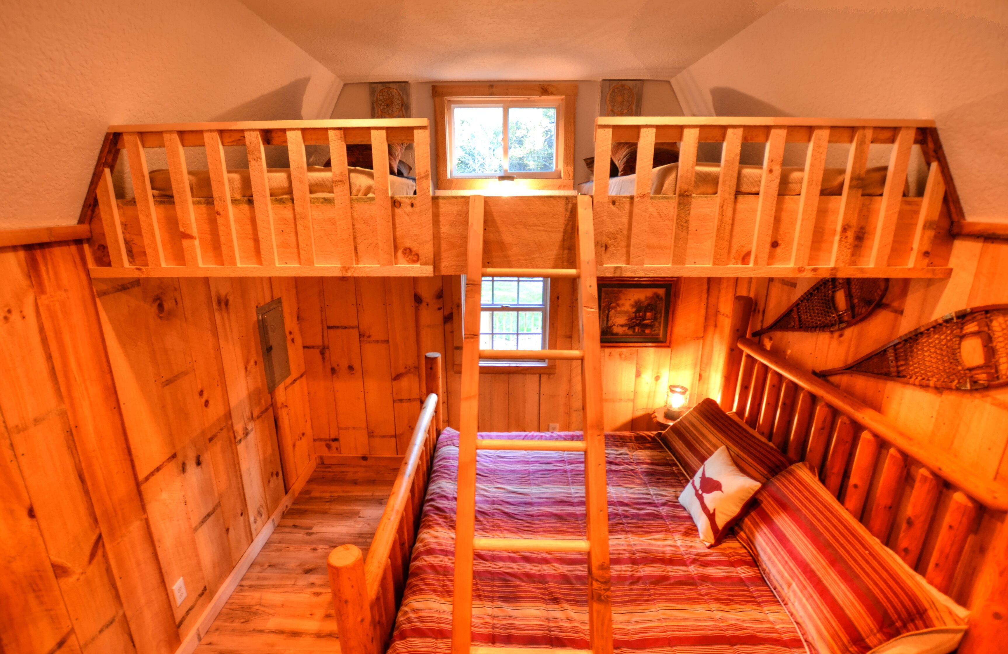 A wooden cabin with a bunk bed and a single bed, featuring a small window and a painting on the wall.