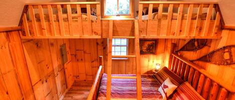 A wooden cabin with a bunk bed and a single bed, featuring a small window and a painting on the wall.