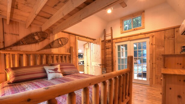 A wooden cabin interior with a bed, a ladder, and a door leading outside. - Cozy Forest Tiny Cabin • Hot Tub • Fire Pit • Pets Welcome • Near NOC & GSMR (Almond)