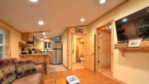 A cozy living room with a sofa, a coffee table, and a TV. There is a kitchen area with a refrigerator and a dining table. - Cozy Forest Tiny Cabin • Hot Tub • Fire Pit • Pets Welcome • Near NOC & GSMR (Almond)