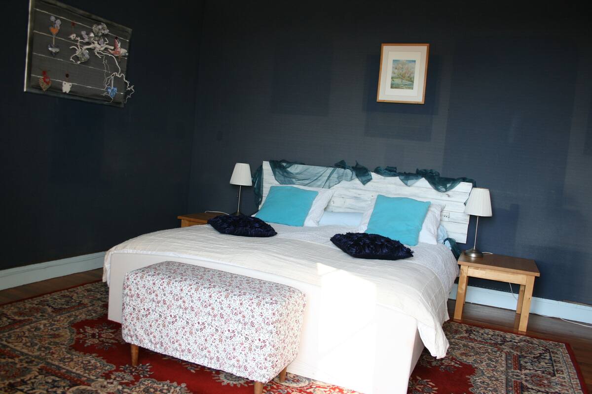A bedroom with a large bed, two bedside tables, a nightstand, a patterned rug, and a framed picture on the wall.
