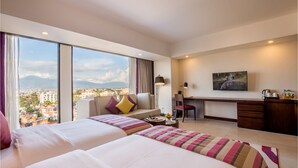 Habitación de hotel con dos camas, un escritorio, una silla, una lámpara de escritorio, un cuadro y vistas a la ciudad.