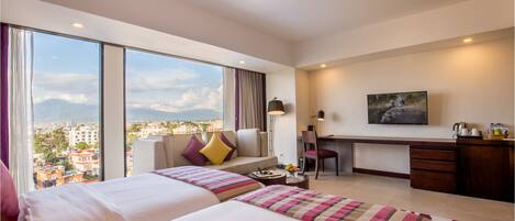 A hotel room with two beds, a desk, a chair, a desk lamp, a painting, and a view of the city.