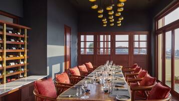 A dining area with a long wooden table set for a meal, flanked by wooden chairs with red cushions. A wine rack with bottles is visible to the left.