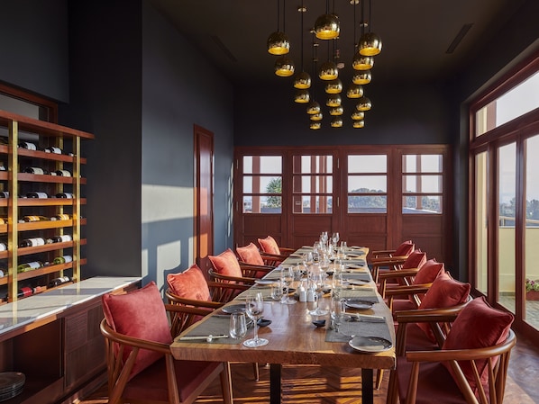 Un comedor con una larga mesa de madera dispuesta para una comida, flanqueada por sillas de madera con cojines rojos. Se ve una vinoteca con botellas a la izquierda.