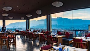 Un restaurante con mesas y sillas, amplias ventanas con vista a la montaña y un paisaje urbano al fondo.