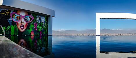 Un mural con fauna y paisaje urbano, una piscina que refleja la escena y una cadena montañosa distante.