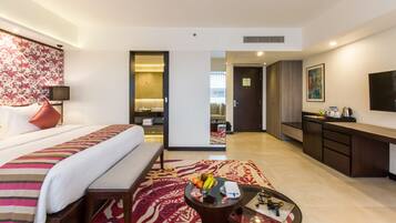 A hotel room with a bed, a bench, a coffee table with fruits, a desk, and a TV.
