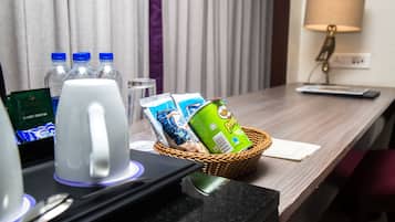 A hotel room desk with a coffee maker, water bottles, a basket with a can of Pringles, a remote control, and a lamp.