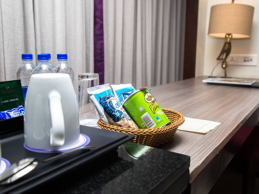 Ein Schreibtisch im Hotelzimmer mit Kaffeemaschine, Wasserflaschen, einem Korb mit einer Dose Pringles, einer Fernbedienung und einer Lampe.