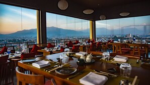 Un restaurante con mesas dispuestas para comer, ventanas amplias que ofrecen vista a la ciudad y a las montañas.