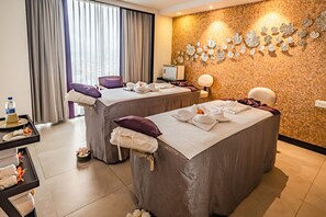 A hotel room with two massage tables, a wall-mounted decorative panel, a small table with towels and a bottle, and a window with curtains. - Vivanta Kathmandu (Lalitpur)