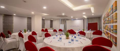 A conference room with round tables, white chairs with red sashes, and a "VIVANTA" sign on the wall.