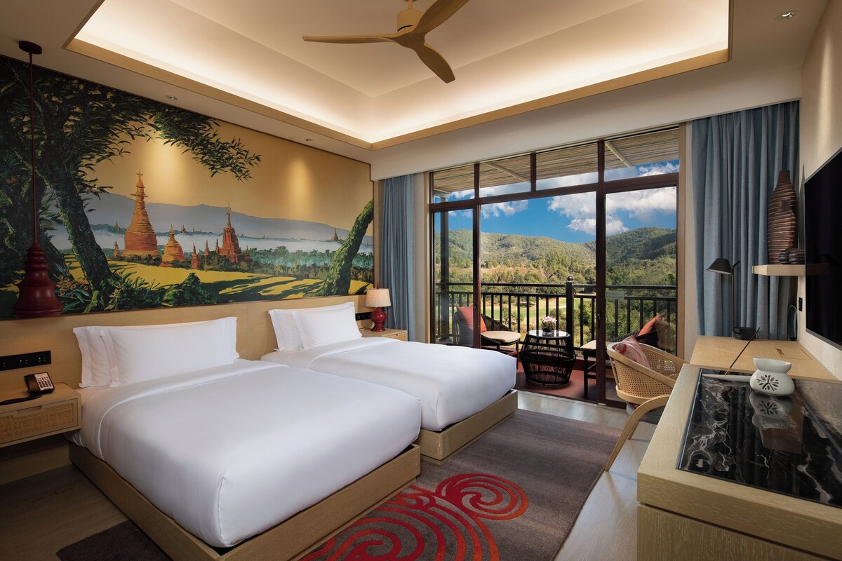 Ein Hotelzimmer mit zwei Betten, einem großen Fenster mit Bergblick, einem Deckenventilator und einem Landschaftsbild.