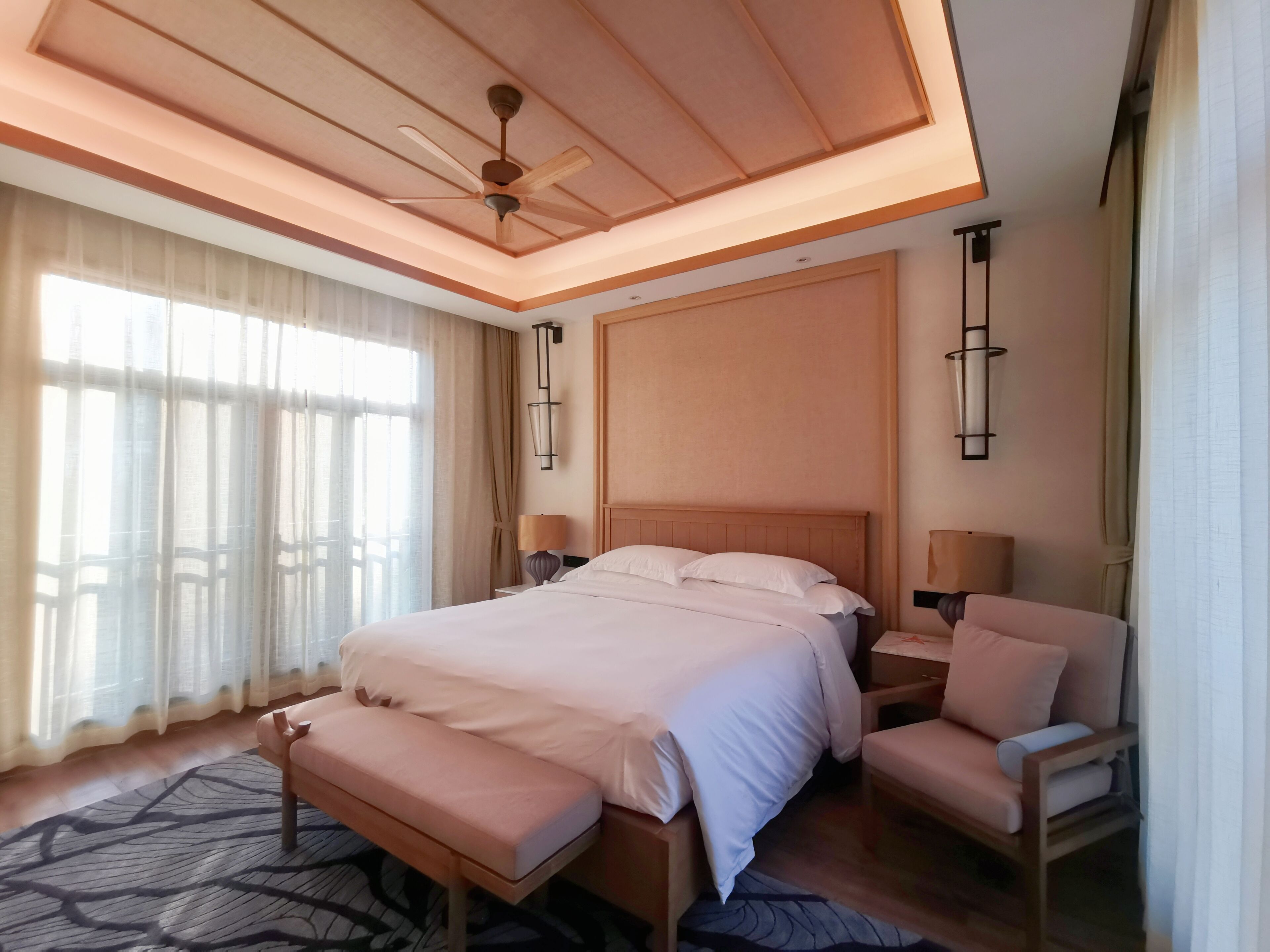 A spacious bedroom with a large bed, a wooden headboard, a ceiling fan, and a view of the outdoors through sheer curtains.
