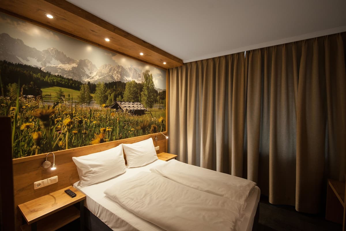 Habitación de hotel con cama, cabecero de madera, mesita de noche y un gran mural en la pared con un paisaje montañoso.