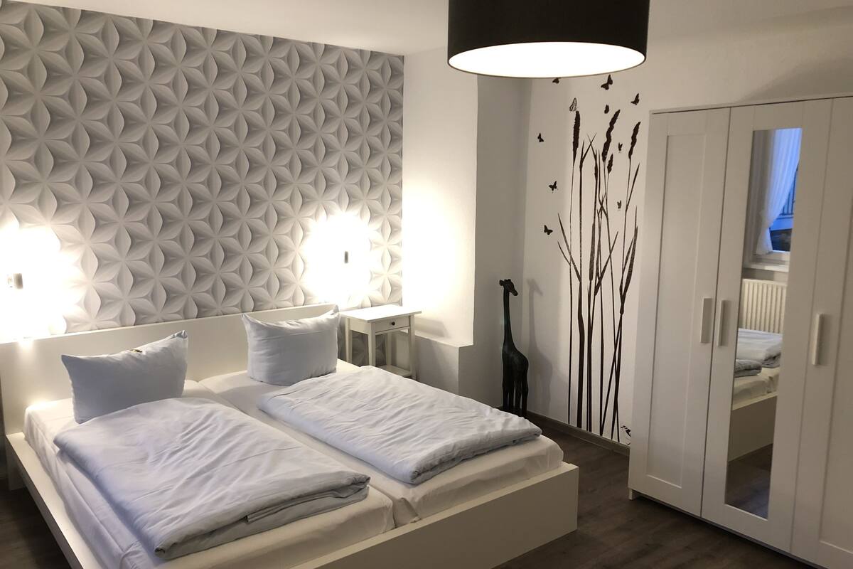 Quarto moderno com uma cama grande, duas mesas de cabeceira, luminária na parede e um guarda-roupa.